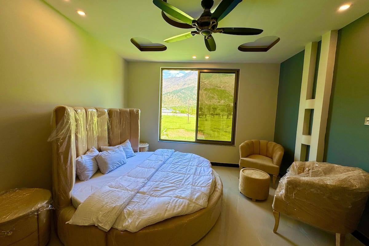 A bedroom with a large bed, a ceiling fan, a window with a view of green fields, and a chair.