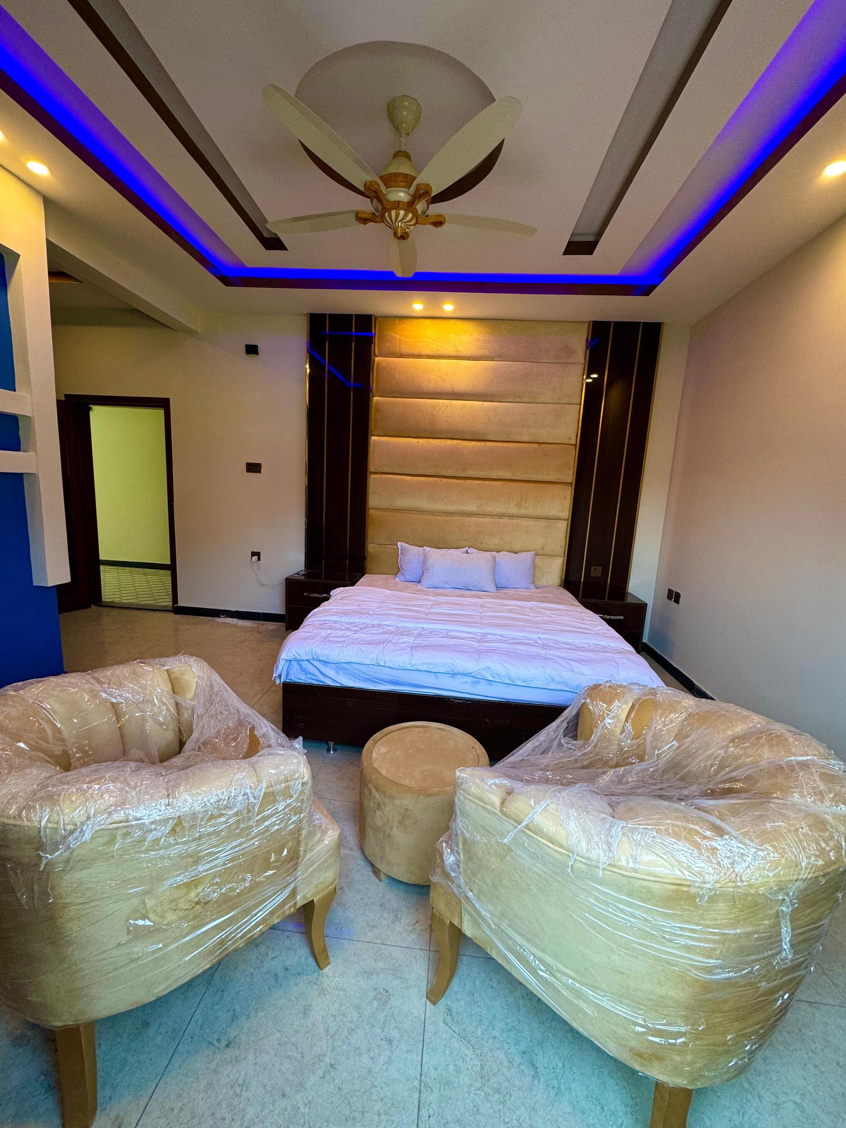 Un dormitorio moderno con una cama grande, dos sillones tapizados, un otoman redondo e un ventilador de techo.