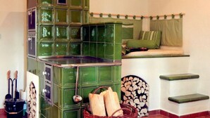Een kamer met een groen betegelde kachel, een houten mand met houtblokken en een bed met groen beddengoed.