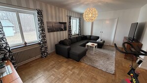 A living room with a sectional sofa, a small table, a TV, a ceiling light fixture, and a radiator.
