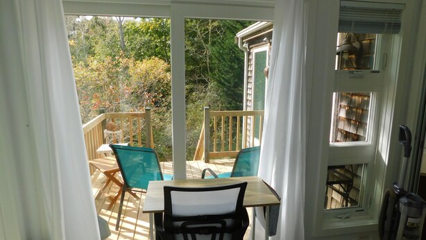 A room with a view of a wooden deck and trees, a table with chairs, and a vacuum cleaner. - Bright and cozy private cottage (CS0037) (Wellfleet)