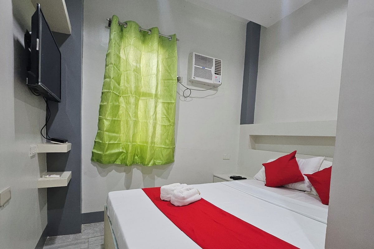 A hotel room with a bed, a television on a shelf, a green curtain, and a white wall.