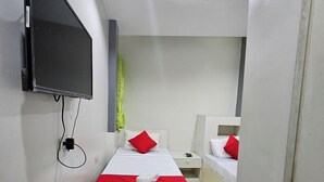 Habitación de hotel con un televisor de pantalla plana grande montado en la pared, dos camas con ropa de cama roja y blanca, una mesita de noche con lámpara, y una mesita de noche pequeña con teléfono.
