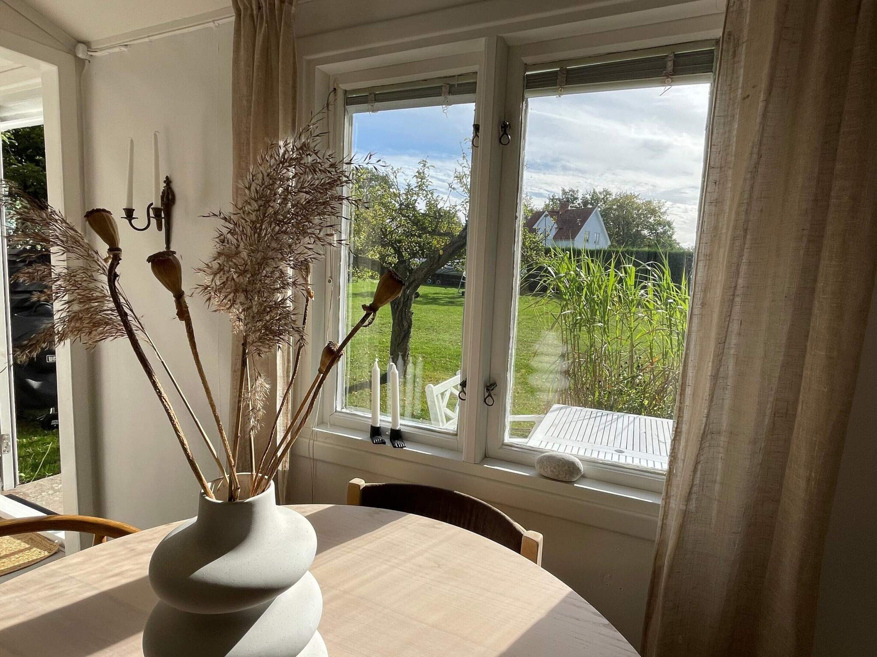 A room with a round table, a vase with dried plants, a window with curtains, and a view of a house and greenery outside.