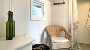 Un bagno con una sedia in vimini, uno scaffale bianco con bottiglie verdi, una finestra e una tenda da doccia.