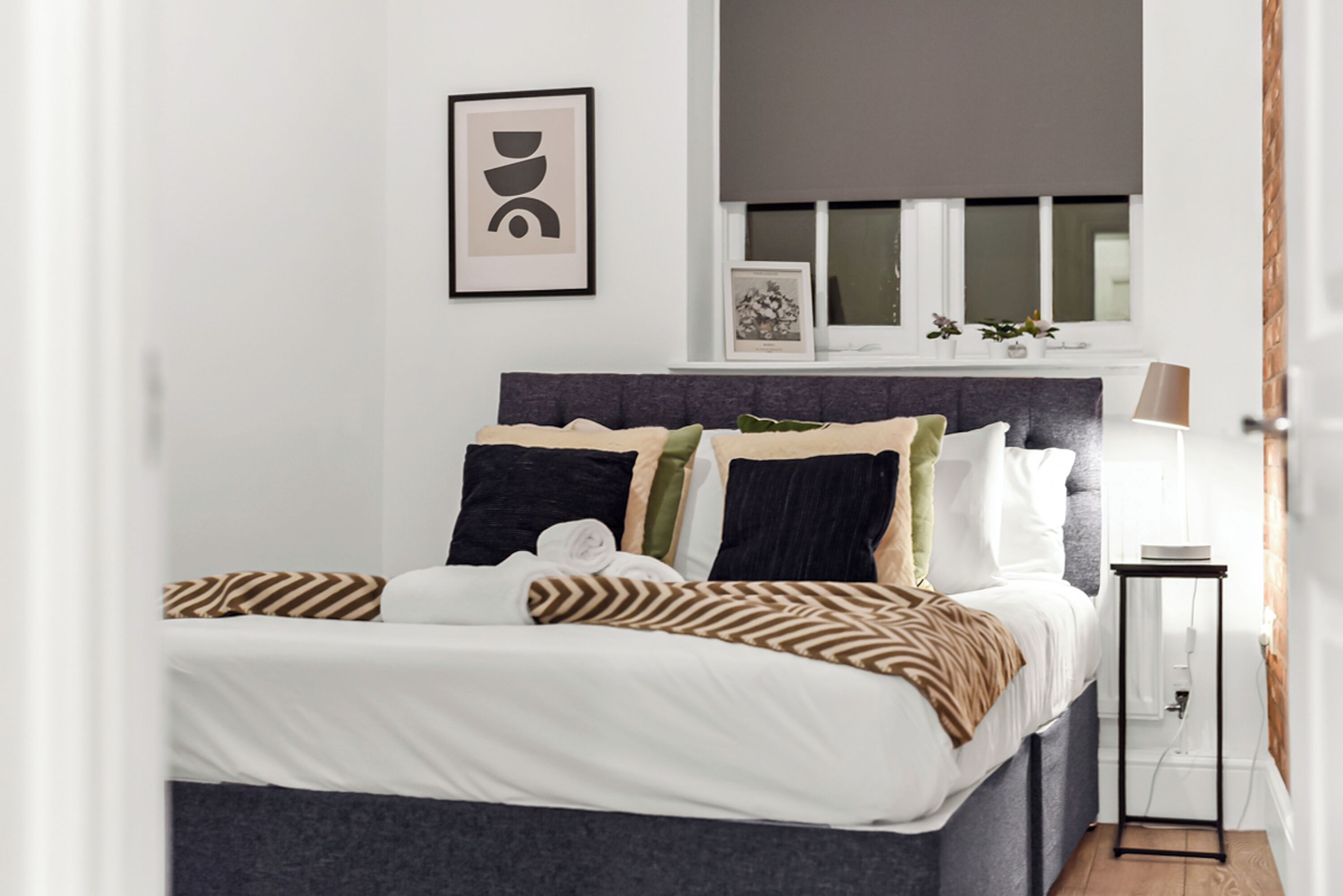 A neatly made bed with a variety of pillows and a blanket, a bedside table with a lamp, and a window with blinds and decorations.