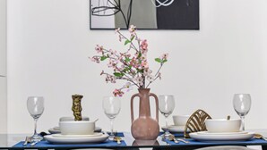 A dining area with a table set for two, featuring a vase with pink flowers, a modern painting, and a blue placemat.