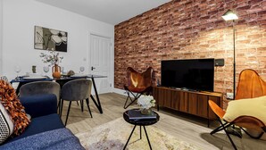 A modern living room with a brick wall, a flat-screen TV, a dining area with chairs and a table, and a blue sofa.