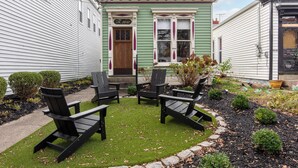 Een groene woning met een voordeur, twee ramen en een klein terras met Adirondack-stoelen en een tafel.