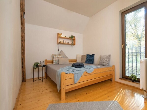 A bedroom with a wooden bed, a small bedside table, a shelf with decorative items, and a window with blinds.