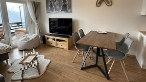 Een moderne woonkamer met een houten eettafel, grijze stoelen, een flatscreen televisie op een houten dressoir, een schilderij van dieren en uitzicht op een berg door een groot raam.