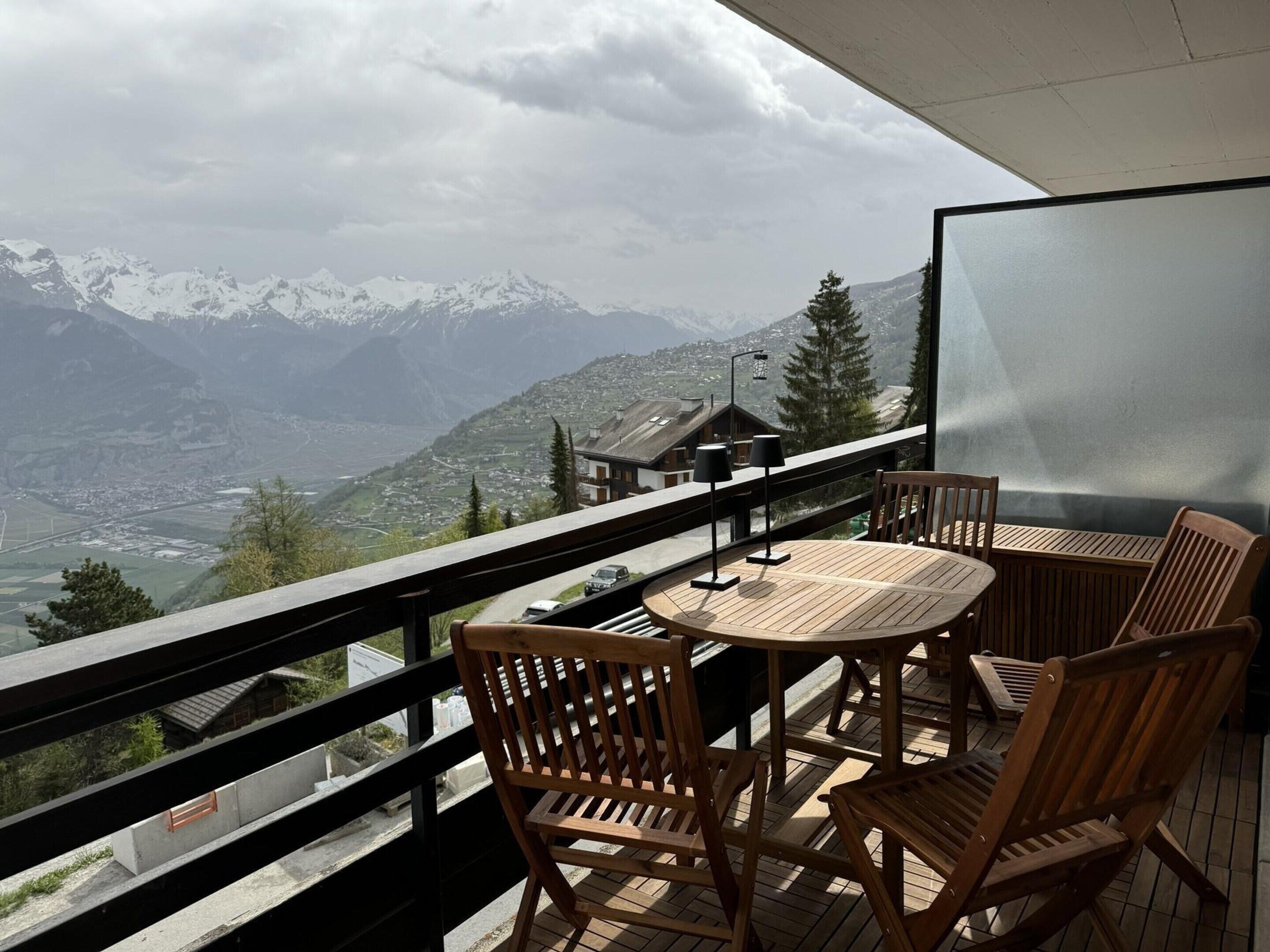 A balcony with a wooden table and chairs, overlooking a mountainous landscape with snow-capped peaks.