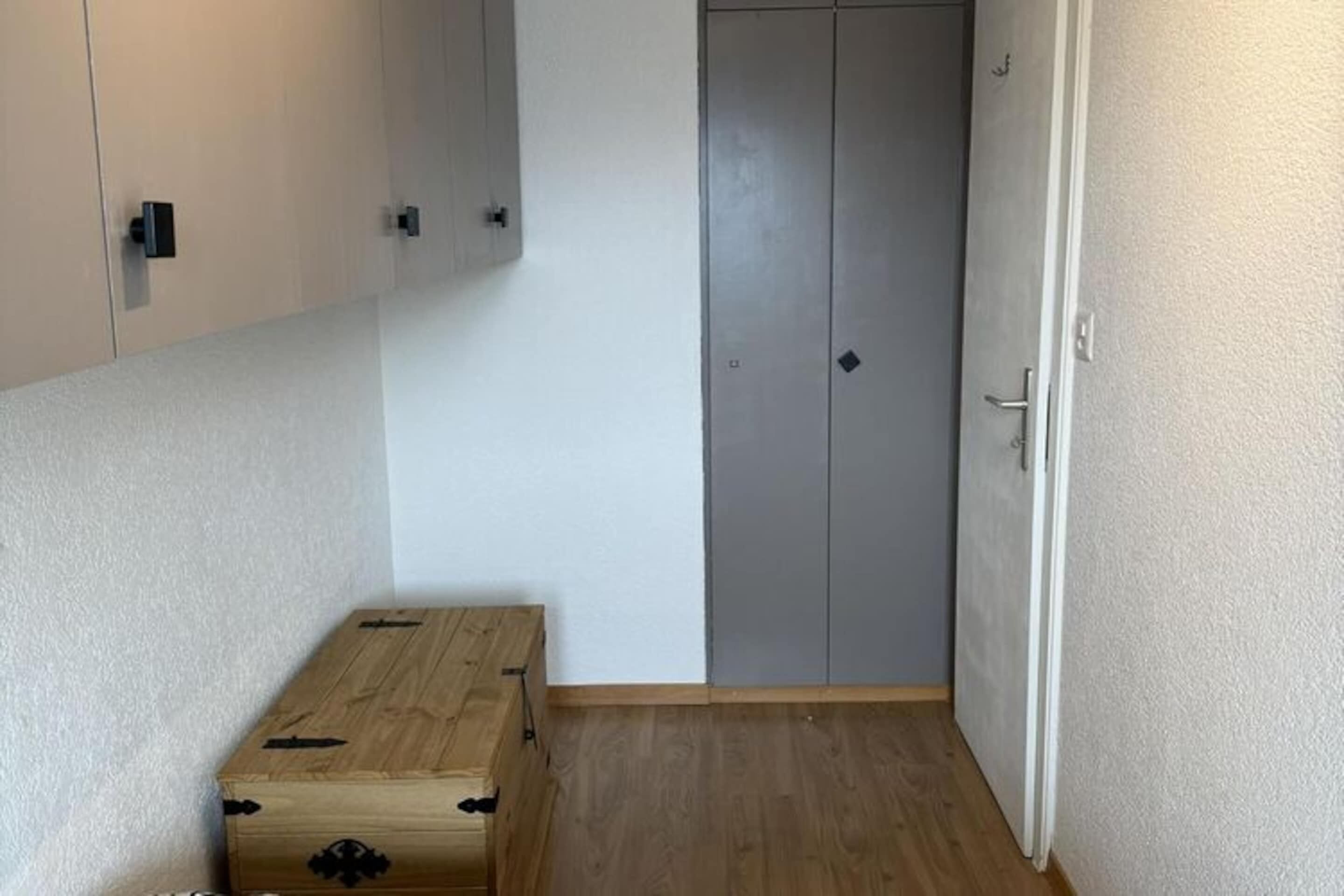 Pieni huone, jossa on puinen arkku, harmaa vaatekaappi ja valkoinen ovi.