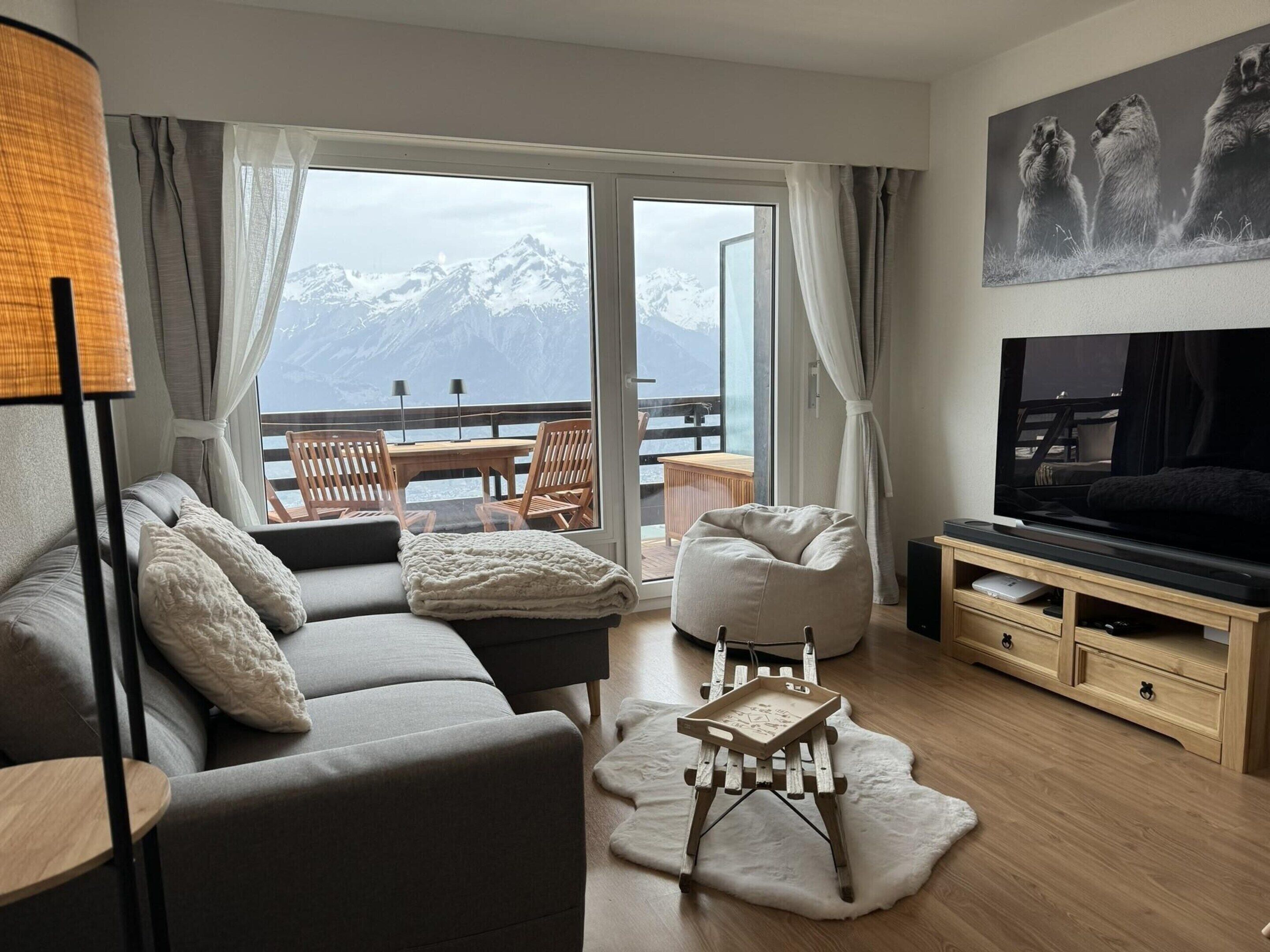 Un salon moderne avec un canapé, une table basse, un téléviseur et une vue sur des montagnes enneigées, visible à travers de grandes fenêtres.