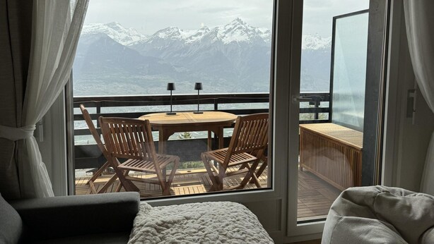 Een kamer met een grote schuifglasdeur die uitkomt op een terras met een houten tafel en stoelen, met uitzicht op een bergachtig landschap.