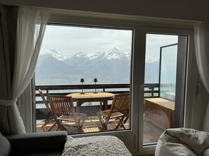 A room with a large sliding glass door leading to an outdoor deck with a wooden table and chairs, overlooking a mountainous landscape. - Lovely Apartment in Valley and Hills (Veysonnaz)
