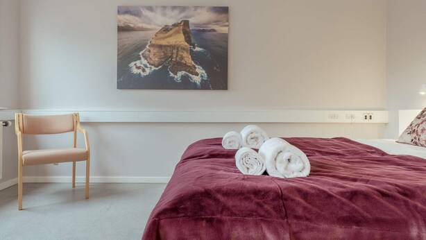 Quarto de hotel com uma cama, uma cadeira, um quadro de paisagem costeira e toalhas dobradas sobre a cama.