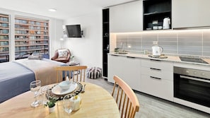 Une cuisine moderne dotée d’un four encastré, un coin repas avec une table préparée pour deux personnes, et une vue sur un immeuble à plusieurs étages à l’extérieur.
