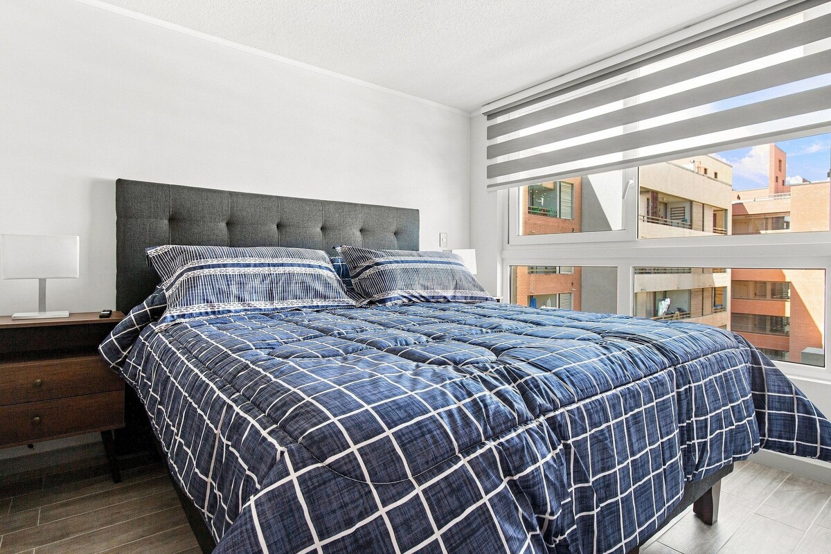 A bedroom with a large bed, a nightstand, a window with blinds, and a view of buildings outside.