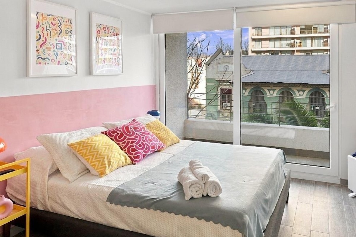 Une chambre à coucher avec un lit, des oreillers, une table de chevet, une fenêtre et des œuvres d’art accrochées au mur.