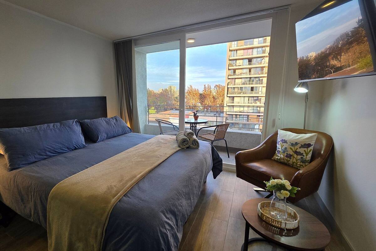Een hotelkamer met een bed, een stoel, een kleine tafel en een balkon met uitzicht op gebouwen en bomen.