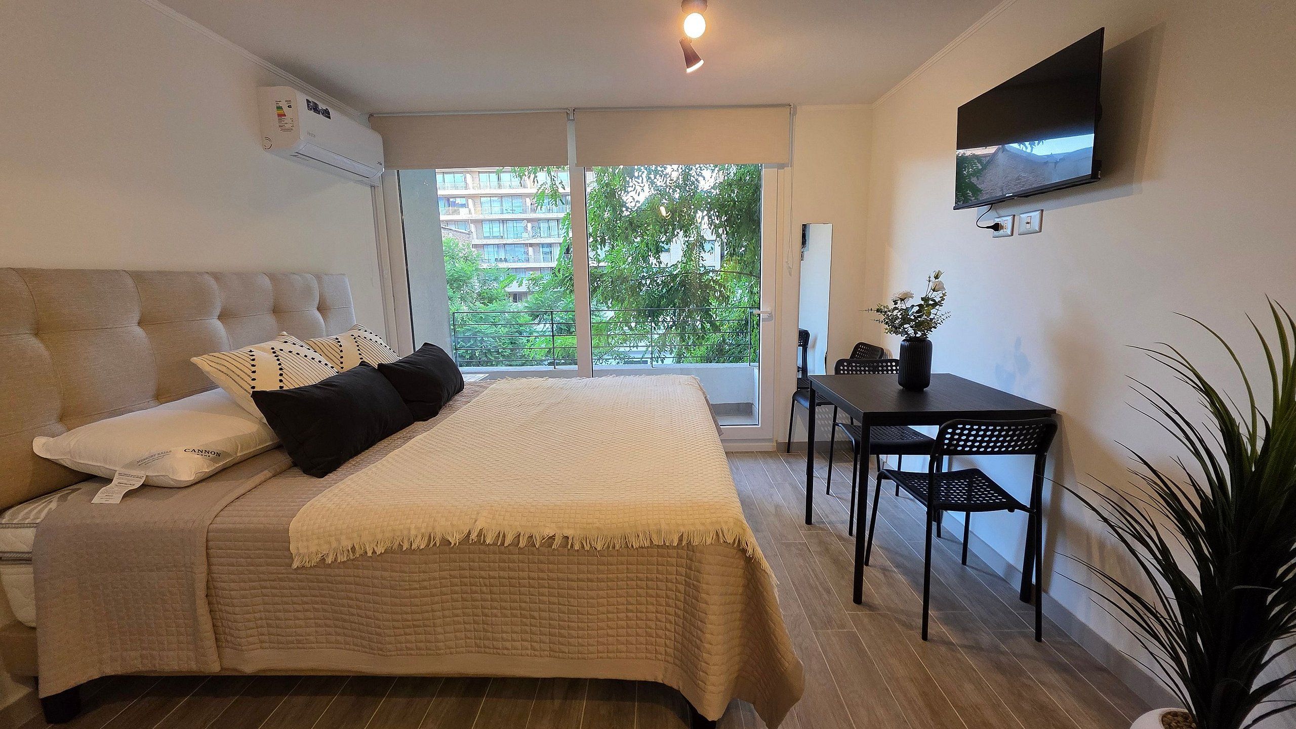 A bedroom with a bed, a desk, a chair, a TV, and a window with a view of buildings and trees.