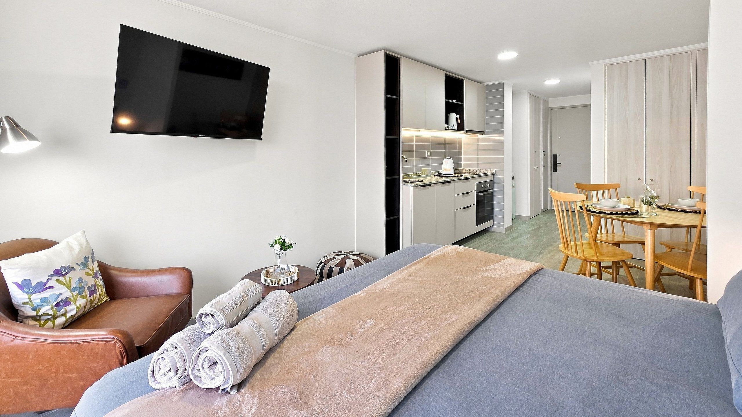 Una camera d'hotel moderna con un divano, una TV a parete, un'area pranzo con tavolo e sedie, e un angolo cottura.