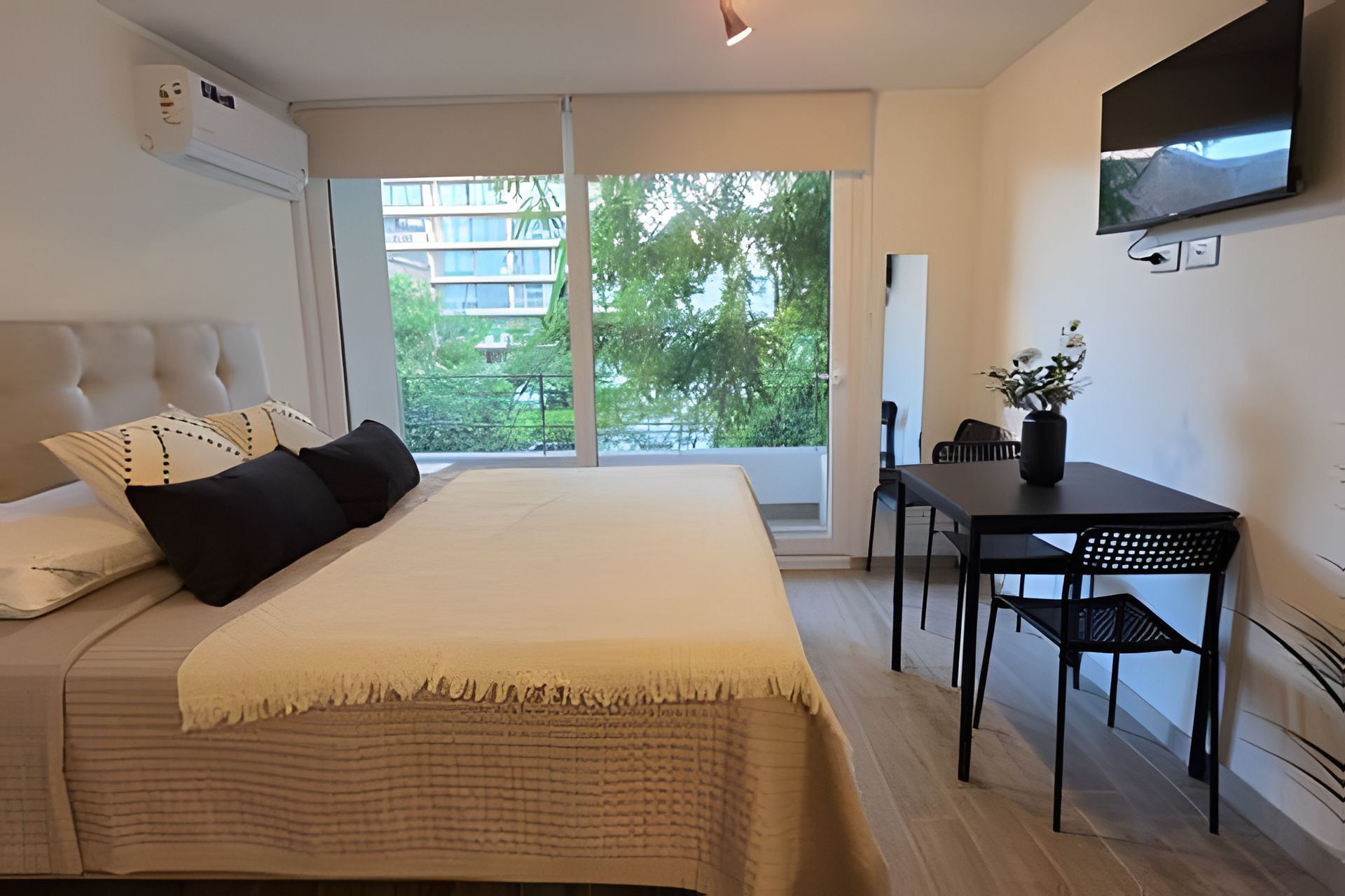 Un dormitorio con cama, escritorio, silla, televisor y ventana con vista a edificios y árboles.