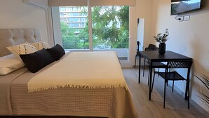 A bedroom with a bed, a desk, a chair, a TV, and a window with a view of buildings and trees. - Oceana Suites Eco España (Santiago)
