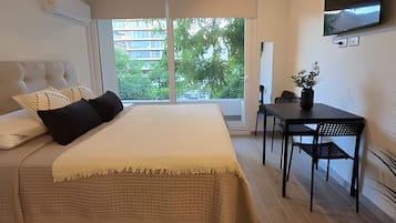 Un dormitorio con cama, escritorio, silla, televisor y ventana con vista a edificios y árboles.