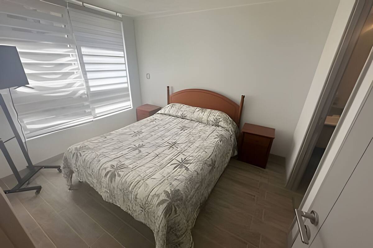 Een slaapkamer met een bed, nachtkastje, lamp en een raam met jaloezieën.