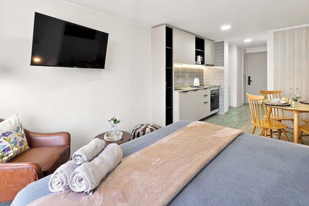 A modern hotel room with a sofa, a TV mounted on the wall, a dining area with a table and chairs, and a kitchenette.