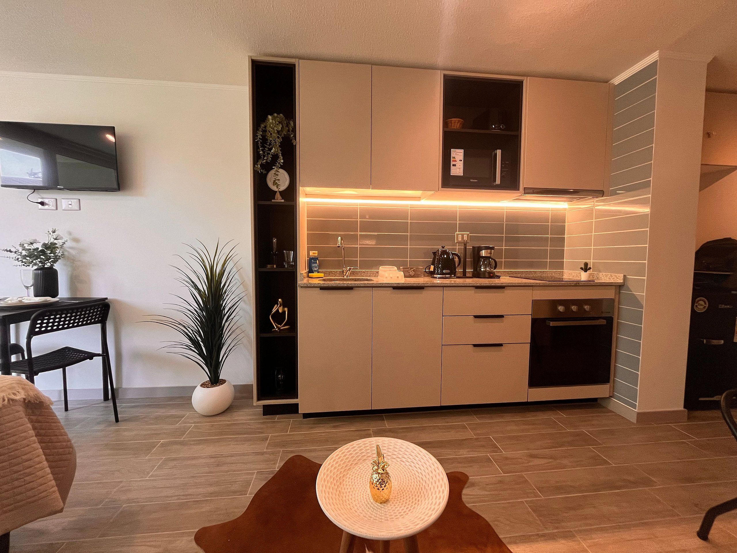 Una cocina moderna con horno empotrado, microondas y cafetera. Hay una zona de comedor con mesa y sillas, y una planta en maceta.