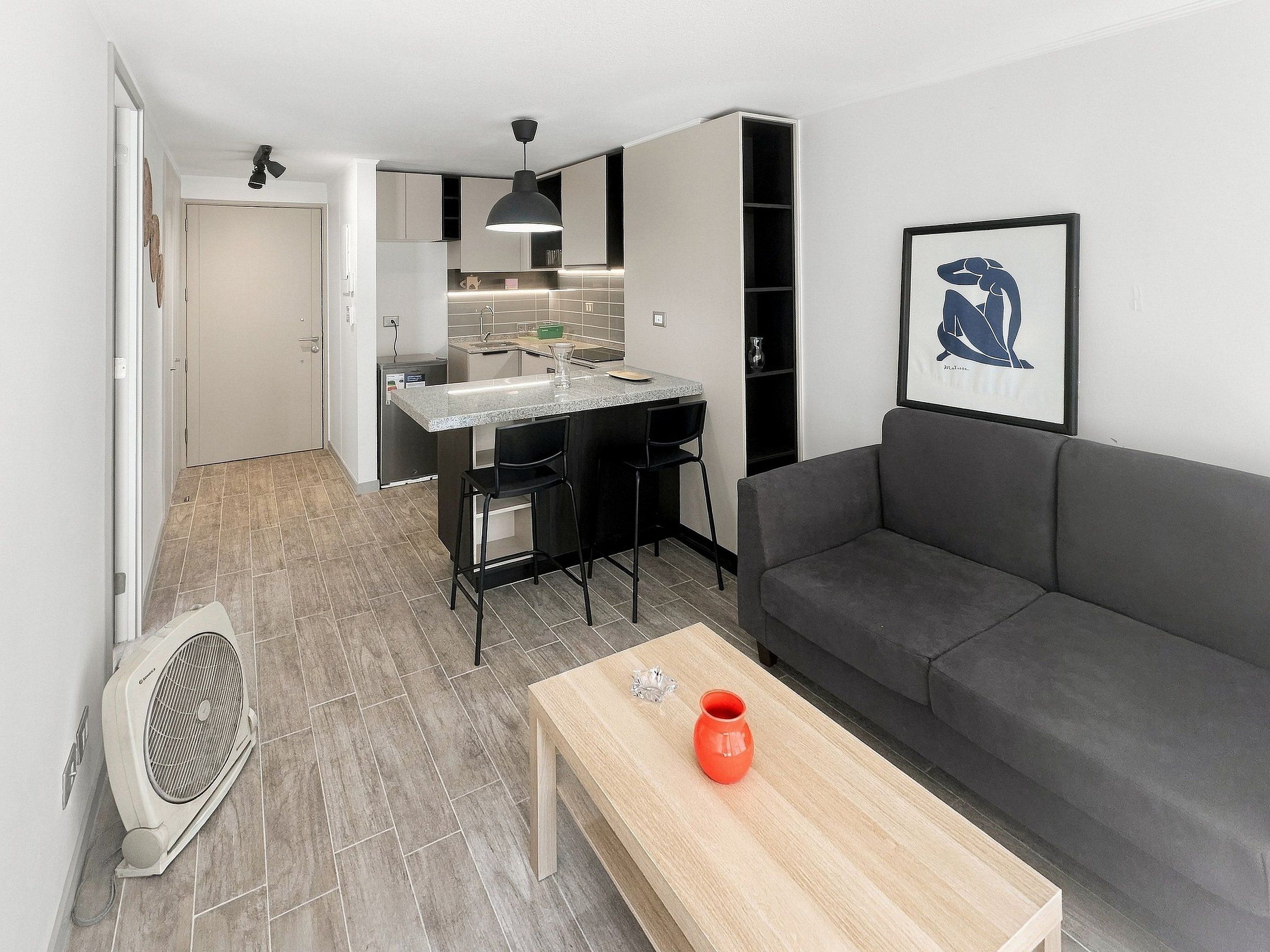 A compact living space with a kitchenette, a grey sofa, a wooden coffee table, and a fan.