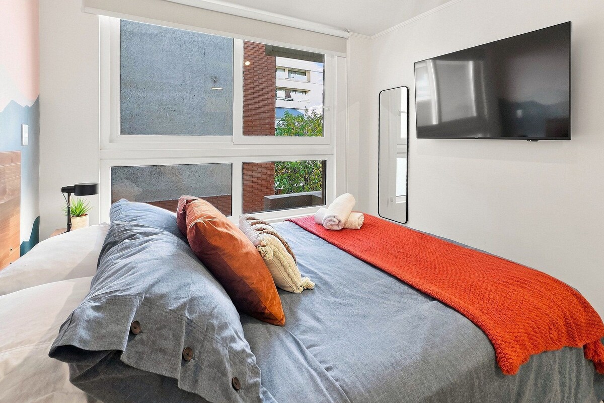 Une chambre moderne avec un lit, une télévision et une fenêtre donnant sur des immeubles.