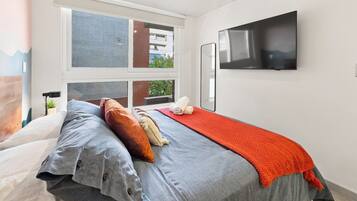Un dormitorio moderno con cama, televisión y ventana con vistas a edificios.