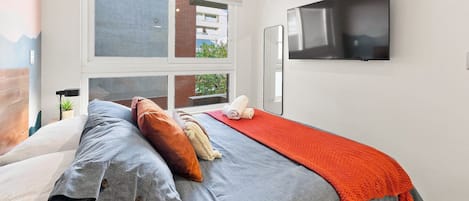 Une chambre moderne avec un lit, une télévision et une fenêtre donnant sur des immeubles.