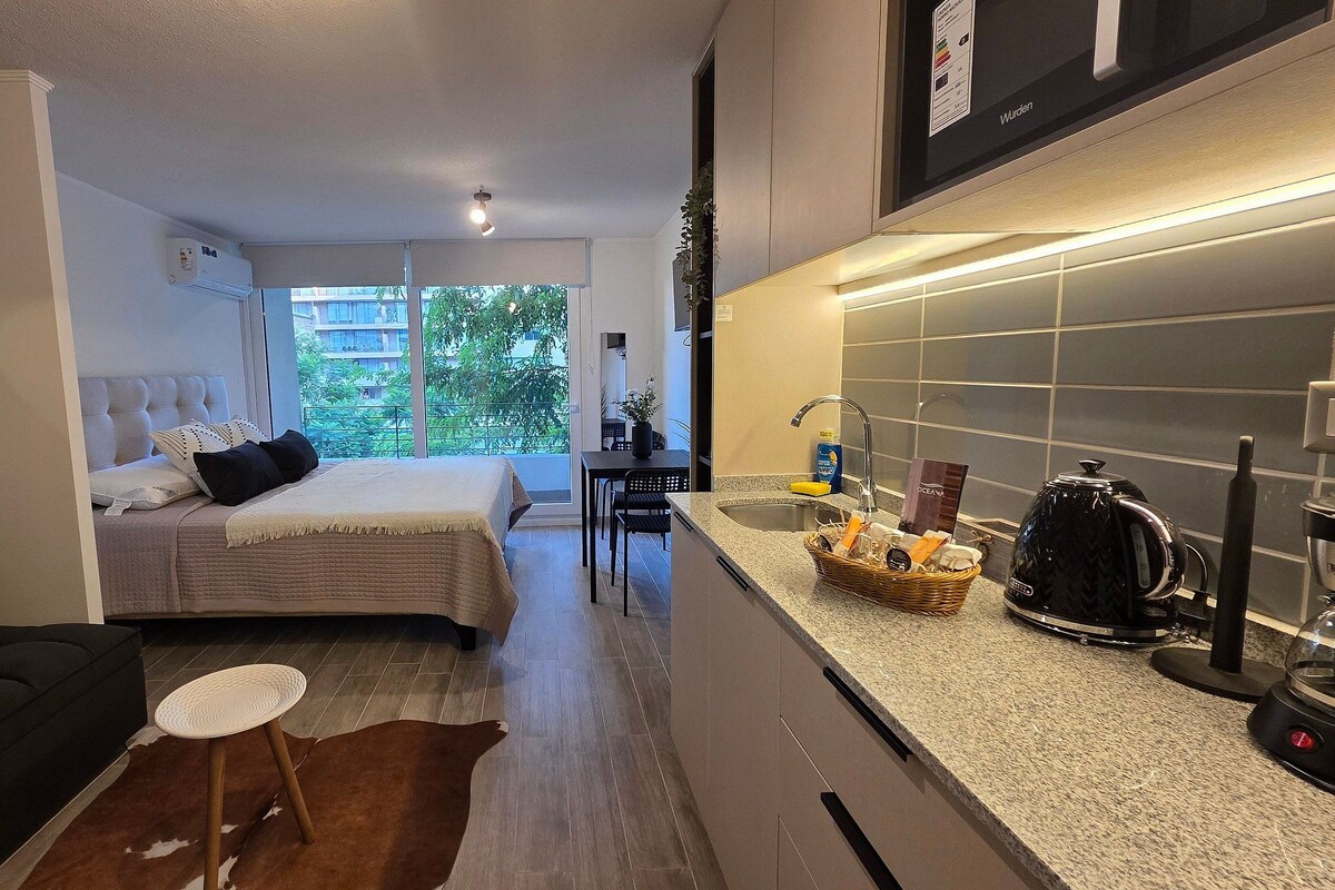 A compact hotel room with a bed, a small sitting area, a kitchenette, and a view of the outdoors.