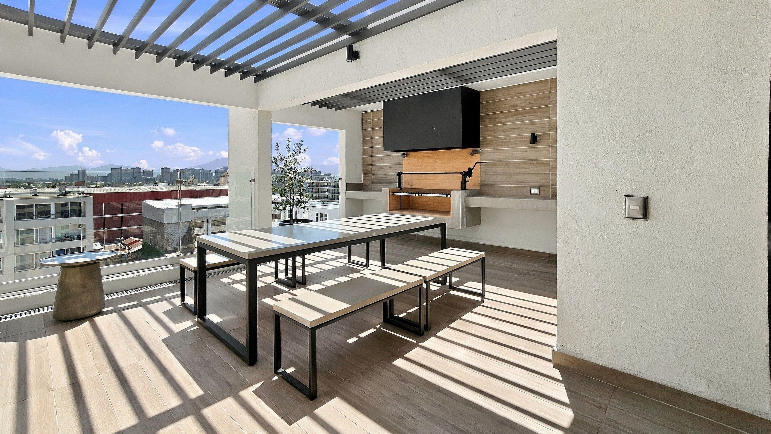 Una terraza al aire libre con una mesa y bancos, un televisor de pantalla plana montado en una pared de madera, y vistas de la ciudad.