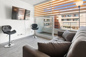 Ein modernes Wohnzimmer mit einer Couch, zwei Sesseln, einem an der Wand befestigten Fernseher und einem großen Fenster mit Jalousien.