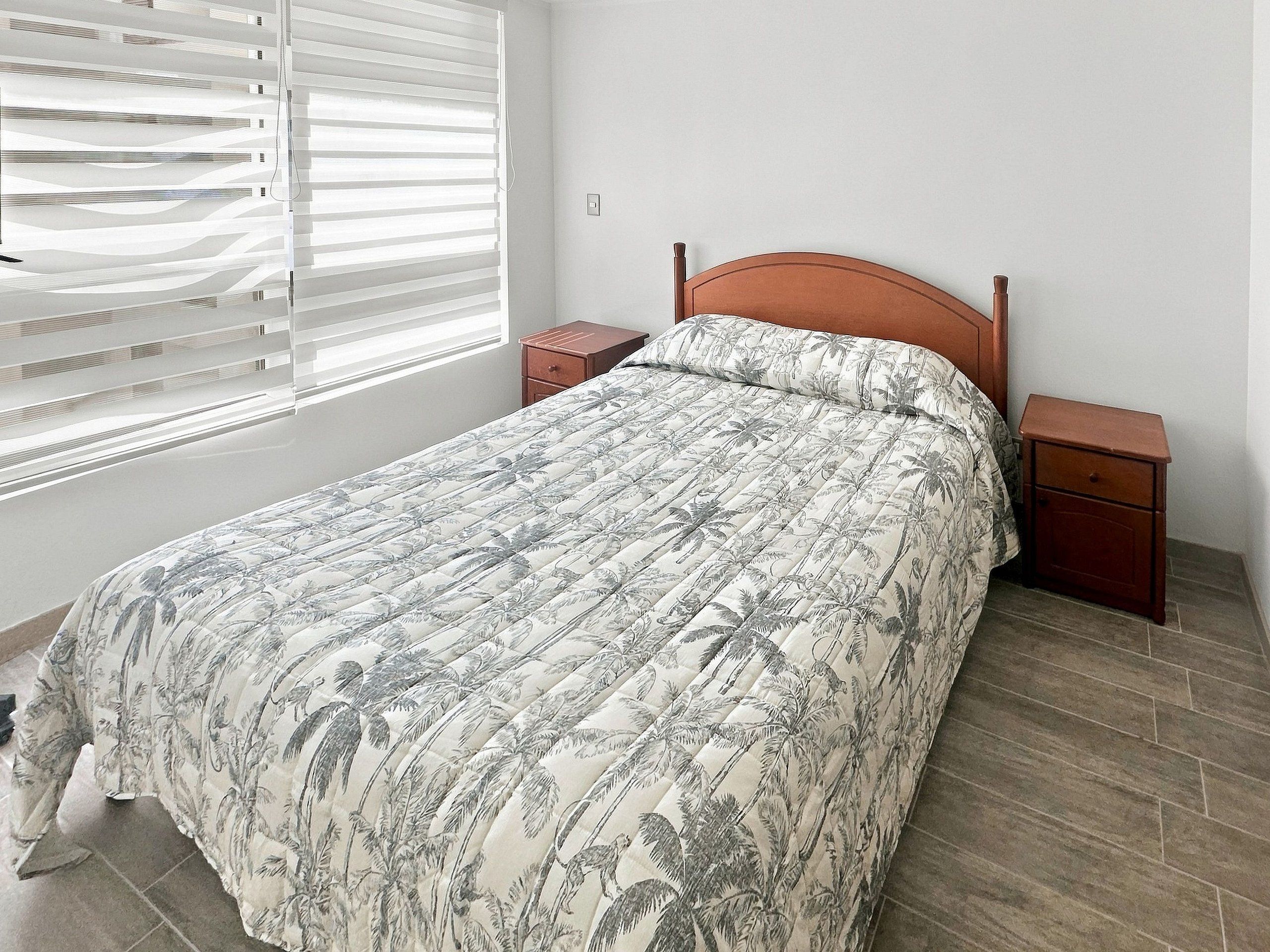 A bedroom with a bed, nightstands, a window with blinds, and a wooden floor.