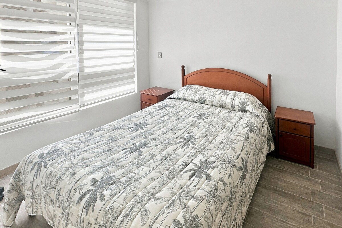 Une chambre à coucher avec un lit, des tables de chevet, une fenêtre avec des stores et un plancher en bois.