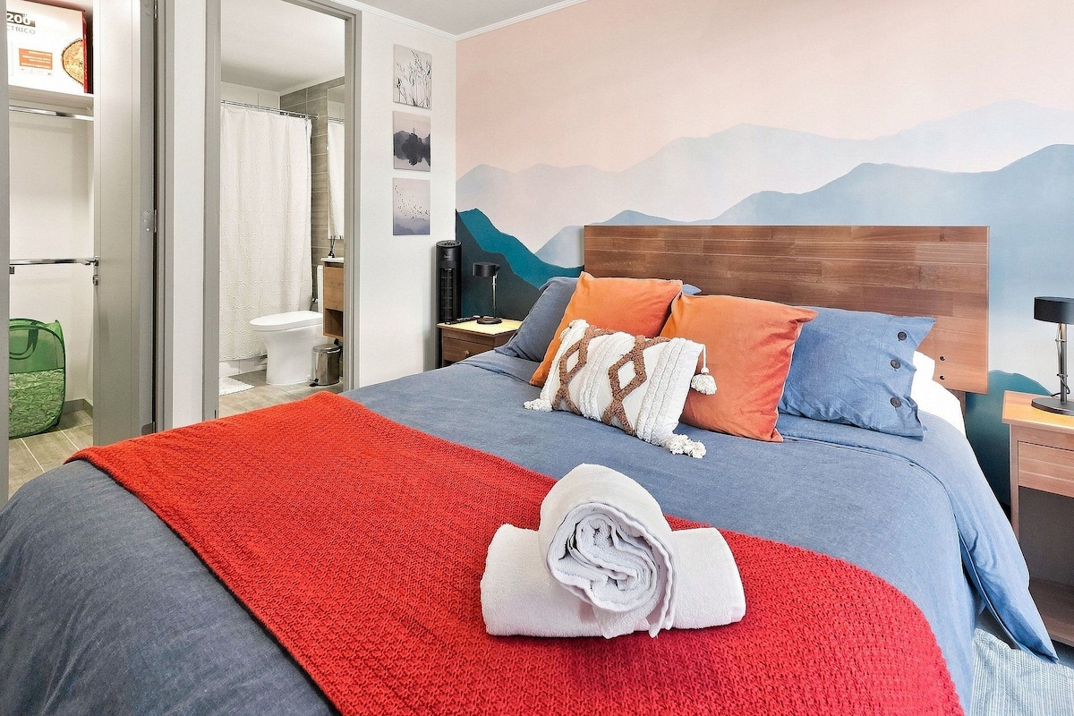 Une chambre d’hôtel comprenant un lit, une table de chevet, un bureau, une chaise, une plante en pot et une salle de bain visible à travers une porte ouverte.