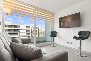 A modern living room with a sofa, two bar stools, a TV, and a large window with blinds. - Oceana Suites Eco España (Santiago)