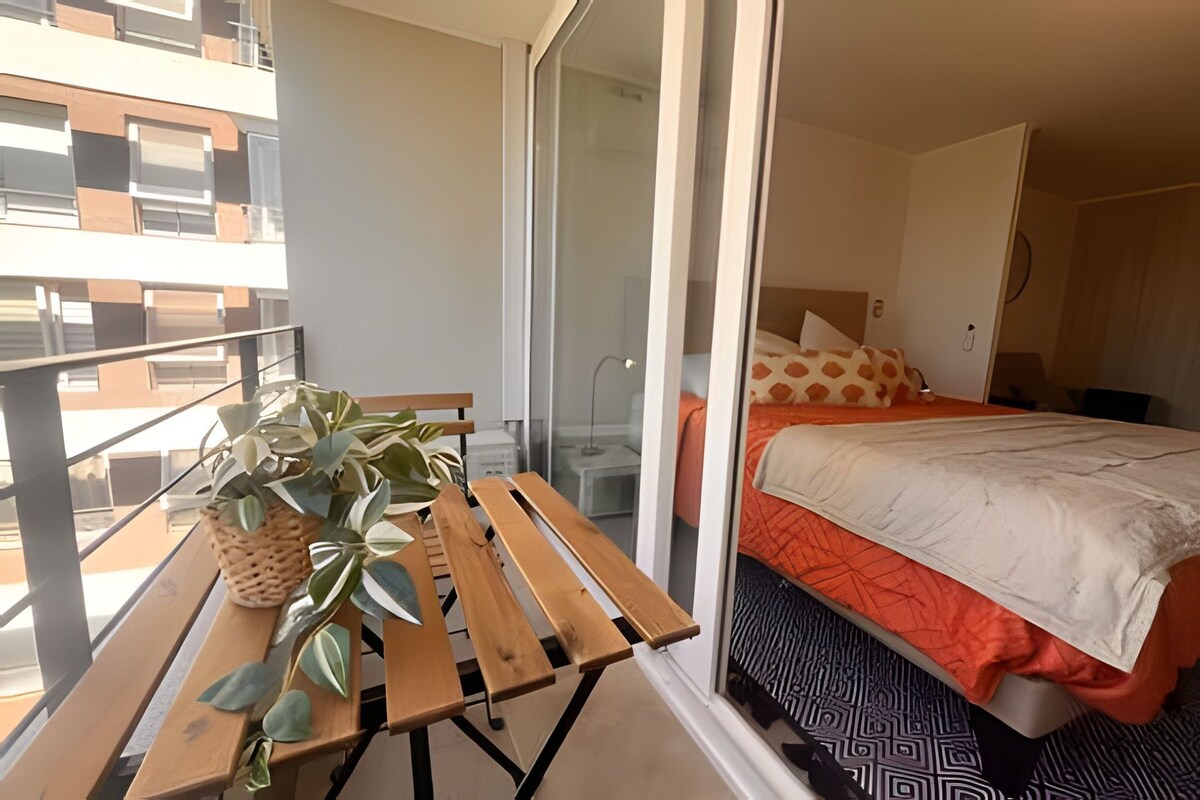 A balcony with a wooden table and potted plants, a bedroom with a bed and patterned bedding, and a view of the building's exterior.