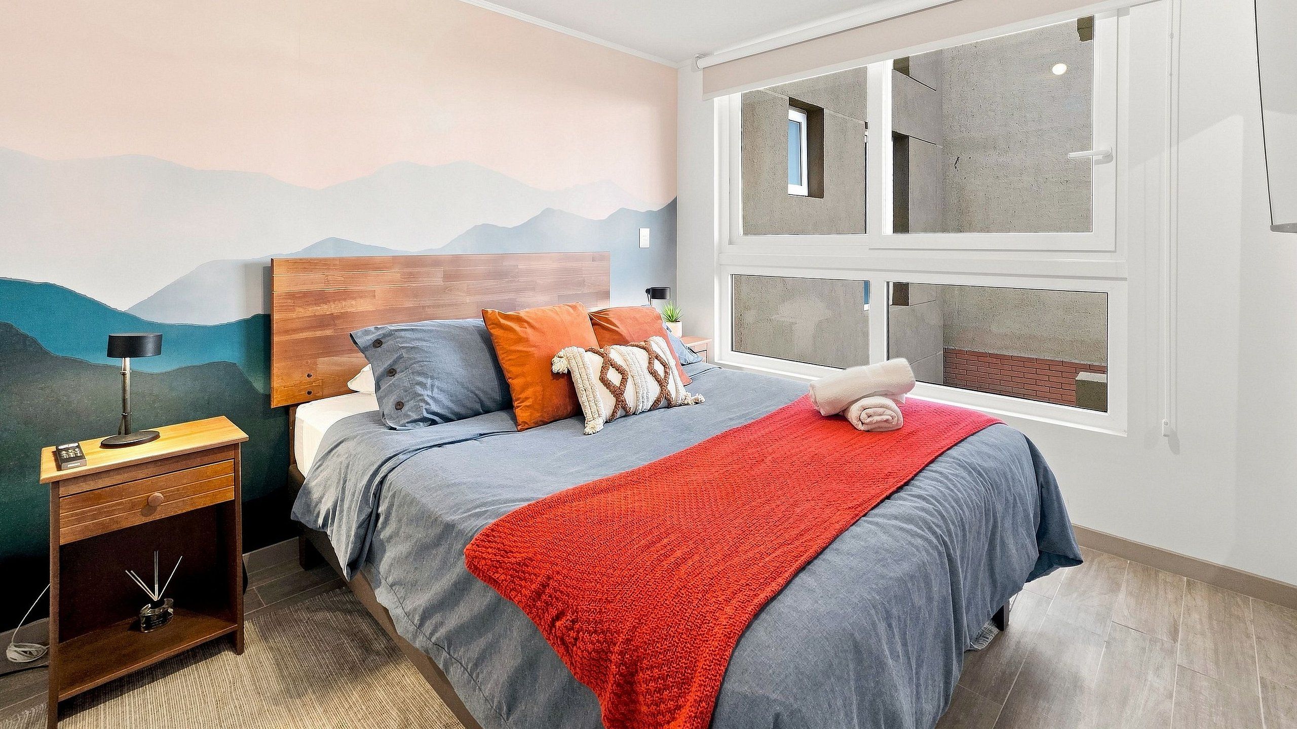 Un dormitorio con cama, mesita de noche, lámpara, ventana con vistas y un mural de montaña en la pared.