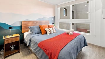 Un dormitorio con cama, mesita de noche, lámpara, ventana con vistas y un mural de montaña en la pared.