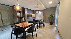 Un espace repas moderne avec des tables en bois et des chaises noires, une cuisine en arrière-plan et une plante en pot.