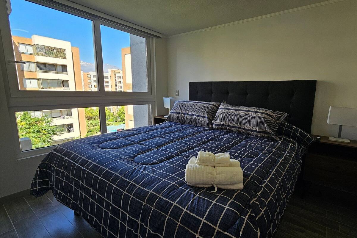 Een slaapkamer met een groot bed, een raam met uitzicht op gebouwen en een nachtkastje met een lamp.