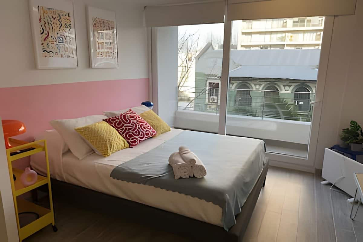 A neatly made bed with pillows and a towel, a small yellow shelf with a toy, and a window with a view of buildings.